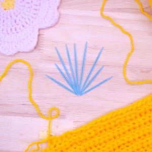 May include: A set of eight light blue plastic yarn needles arranged in a fan shape, positioned between a light pink crocheted flower with a yellow centre and a piece of bright yellow crocheted fabric with loose yellow yarn strands.