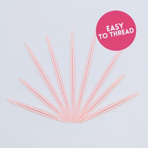 May include: A collection of pale pink yarn needles arranged in a fan shape. The needles are made of plastic and have a pointed end and a slit for threading yarn. A pink circle with the words "EASY TO THREAD" is in the upper right corner.