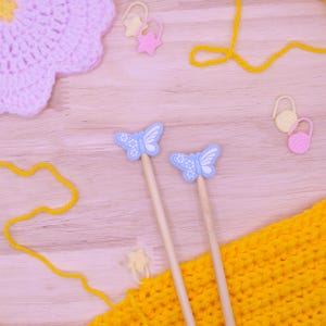 May include: Two bamboo knitting needles featuring light blue butterfly-shaped toppers with white floral details. The needles are accompanied by yellow yarn, a light pink crocheted flower, and several small plastic stitch markers in star and ball shapes.
