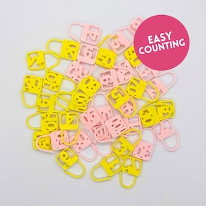 Könnte beinhalten: Eine Ansammlung gelber und rosa Maschenmarkierer, jeweils mit einer ausgeschnittenen Zahl. Die Markierungen haben die Form eines Vorhängeschlosses. Ein rosa Kreis mit dem Text "EASY COUNTING" befindet sich in der oberen rechten Ecke.