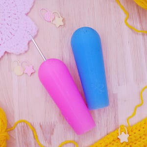 May include: A bright pink crochet hook with a thick, ergonomic handle and a metallic tip, positioned next to a solid blue ergonomic handle. The items are surrounded by yellow yarn, a pink crocheted piece, and small star-shaped stitch markers.