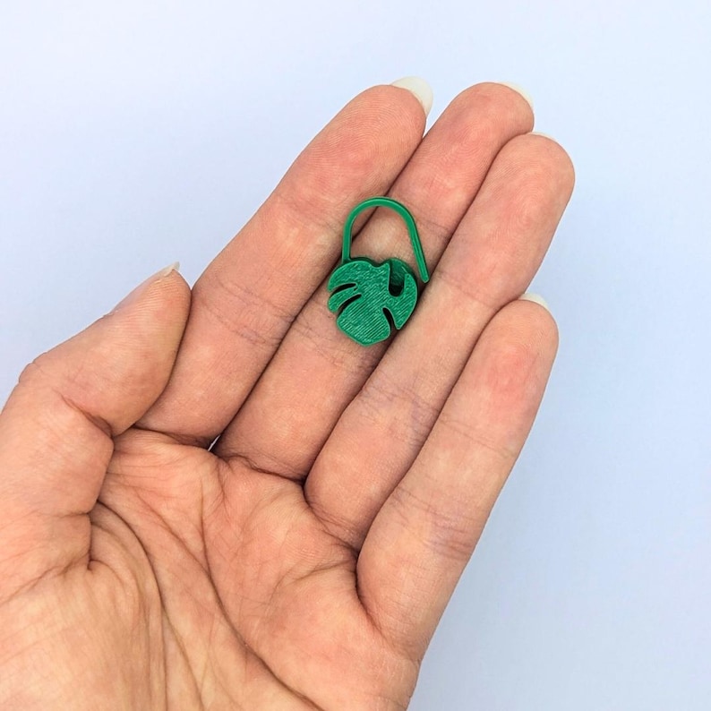 Puede incluir: Un peque&ntilde;o dije verde en forma de hoja de Monstera con un lazo. El dije se sostiene en la mano de una persona sobre un fondo blanco. El dije es de un tono verde vibrante.