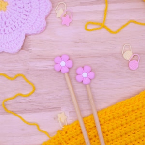 May include: Two wooden knitting needles with light pink flower-shaped toppers, accompanied by a yellow crocheted fabric piece, yellow yarn, a light pink crocheted flower, and several small plastic stitch markers in star and yarn ball shapes.