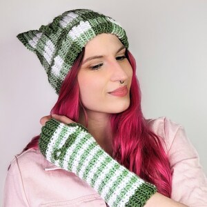 Könnte beinhalten: Eine grün-weiß gestreifte Strickmütze mit spitz zulaufenden Ohren. Die Mütze wird von einer Person mit pinkfarbenem Haar getragen. Die Person trägt auch einen grün-weiß gestreiften Fingerhandschuh.