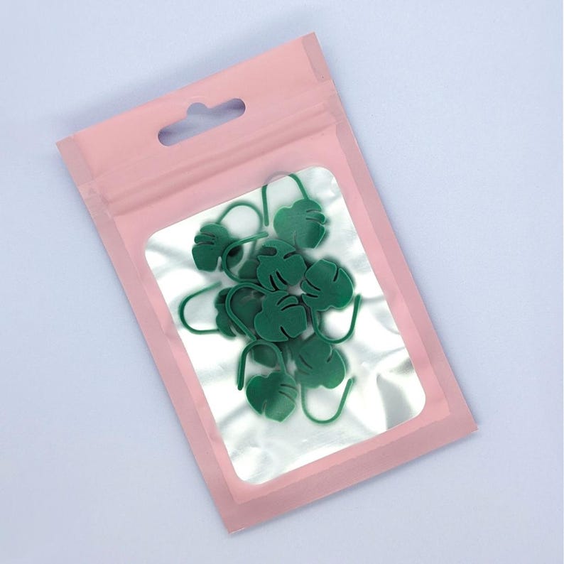 Puede incluir: Una bolsa de pl&aacute;stico transparente con borde rosa que contiene varios marcadores de puntada verdes con forma de hoja. Los marcadores de puntada est&aacute;n hechos de un material suave y tienen un gancho en la parte inferior. La bolsa tiene un peque&ntilde;o agujero en la parte superior para colgar.