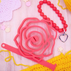 Könnte beinhalten: Ein rotes rosenförmiges Bastelwerkzeug mit einem Schlitz, ein rotes Perlenarmband mit Herzanhänger und gelbes Garn sind auf einer hellen Holzoberfläche angeordnet. Rosa und gelbe Bastelmaterialien sind verstreut.