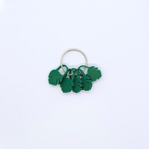 Puede incluir: Un conjunto de colgantes verdes con forma de hoja unidos a un llavero de metal plateado. Los colgantes son de color verde vibrante y tienen patrones detallados de venas de hojas. El llavero es circular y mantiene unidos los colgantes.