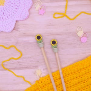 May include: Two wooden knitting needles with sunflower-shaped toppers in small pots, accompanied by a yellow crocheted fabric piece, a light pink crocheted flower, yellow yarn, and several plastic stitch markers in star and yarn ball shapes.