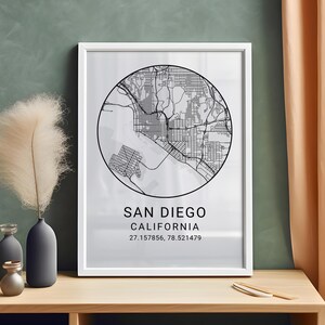 Puede incluir: Un cuadro enmarcado con un mapa en blanco y negro de San Diego, California, con el nombre de la ciudad y sus coordenadas. El mapa es circular, sobre un fondo blanco, y está dentro de un marco blanco. El cuadro se muestra sobre una superficie de madera.
