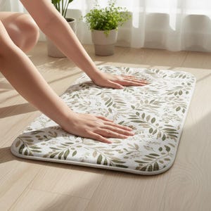 Puede incluir: Una alfombra de baño rectangular con fondo blanco y un estampado de hojas y ramas verde oliva. La alfombra tiene esquinas redondeadas y un borde blanco. Está colocada sobre un suelo de madera clara.