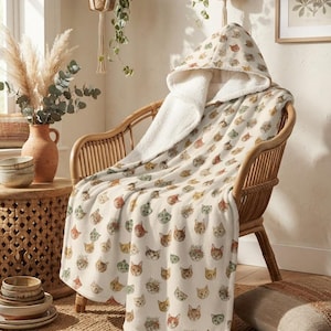 Puede incluir: Una manta con capucha que presenta un estampado integral de varias caras de gatos en tonos naranja, marrón, gris y blanco. La manta tiene un forro de forro polar sherpa de color crema y está colocada sobre una silla de madera curva.