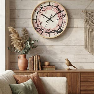 Puede incluir: Un reloj de pared redondo con marco de madera clara que presenta un círculo central color crema rodeado por un anillo rosa. La esfera muestra ramas negras con flores de cerezo rosas, manecillas negras y un segundero fino de color negro.