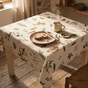 Puede incluir: Un mantel cuadrado con un estampado botánico de limones, aceitunas y ramitas sobre un fondo crema. La mesa está puesta con un plato de cerámica que contiene una rebanada de pan, un cuchillo de plata, una cuchara pequeña y una taza humeante.