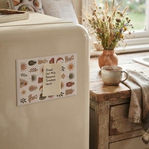 Puede incluir: Una lámina magnética rectangular con un patrón repetitivo de soles, jarrones y hojas adherida a un refrigerador de color crema. Una nota adhesiva de color amarillo pálido con el texto escrito a mano Bread, Oat Milk, Bananas, Tomatoes, Herbs está pegada en el centro.