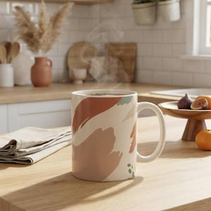 Puede incluir: Una taza de cerámica con base color crema que presenta patrones de pinceladas abstractas en tonos terracota, rosa apagado y verde salvia. La taza tiene un asa curva y contiene una bebida caliente humeante sobre una superficie de madera.