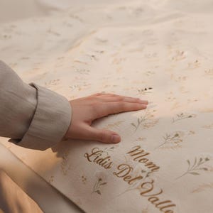 Puede incluir: Manta crema con estampado floral y el texto bordado "Mom Lidis Del Valle". La manta tiene un aspecto suave y texturizado y está adornada con delicados diseños botánicos en tonos beige y marrón.