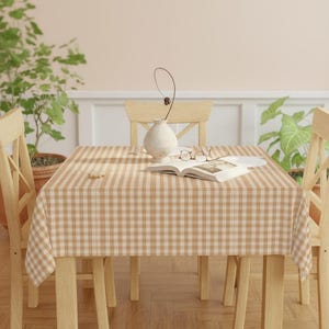 Puede incluir: Una mesa de comedor cuadrada cubierta con un mantel a cuadros beige y blanco. Un jarrón con un tallo decorativo, un libro abierto y gafas están sobre la mesa. Se ven dos sillas de madera, junto con plantas en macetas.