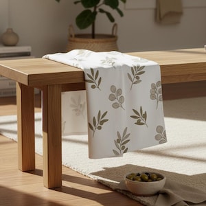 Puede incluir: Un camino de mesa blanco con un patrón repetido de ramas de olivo en tonos verdes y marrones. El camino de mesa está colocado sobre un banco de madera clara. Un pequeño cuenco de aceitunas está sobre una alfombra beige.