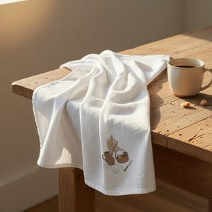 Puede incluir: Toalla de cocina de lino blanco con una ilustración bordada de una jarra, una cesta y trigo. Una taza de café está sobre una mesa de madera, con unos cubitos de azúcar. La toalla está sobre la mesa.