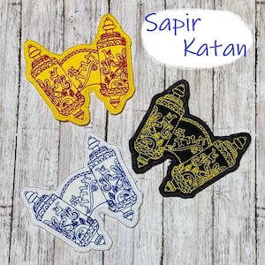 May include: Three embroidered patches in yellow, black, and blue, each featuring a stylized lantern design with intricate floral patterns. The patches are outlined in white and rest on a wooden surface. The text "Sapir Katan" is in a blue bubble.