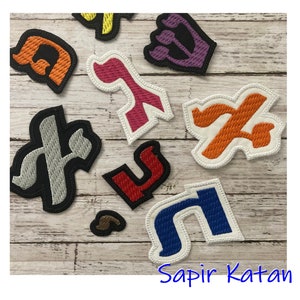 May include: A set of nine embroidered felt patches in various colours, each featuring a different Hebrew letter. The letters are arranged on a light wood background. The text "Sapir Katan" is visible at the bottom of the image.
