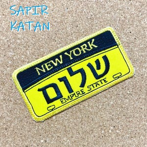 Puede incluir: Parche bordado amarillo y negro con el texto "NEW YORK" y "EMPIRE STATE" y la palabra hebrea "שלום" en negro.