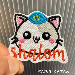 May include: Embroidered patch featuring a cartoon cat with a blue yarmulke and a Star of David. The word "Shalom" is written in orange script below the cat's face. The patch has a white background and black outline.