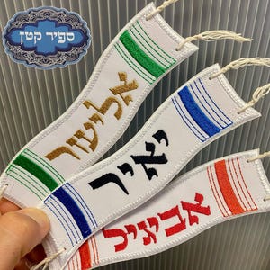 May include: Three rectangular white fabric bookmarks with embroidered Hebrew text in gold, black, and red. Each bookmark features colored stripes in green, blue, or orange at the ends and includes a small white tassel string at the top corner.