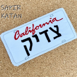 May include: A white rectangular patch with a light blue border, resembling a California license plate. The word "California" is embroidered in red script, and black Hebrew letters are below. The text "SAPIR KATAN" is above.