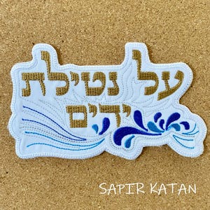 May include: A white fabric patch with blue and gold embroidery. The patch features the Hebrew words "עַל נְשִׁיאַת" and "דְּרָכִים" in gold thread, along with a blue floral design. The words "SAPIR KATAN" are printed in black below the patch.