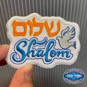 以下が含まれることがあります： ヘブライ語と英語で「Shalom」という言葉が刺繍されたパッチ。鳩が描かれています。パッチは白い縁取りがあり、手で持たれています。ヘブライ語はオレンジ色、英語は青色です。