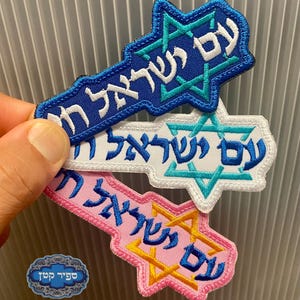 Pode incluir: Três emblemas bordados em forma de Estrela de Davi com uma cauda. O emblema superior é azul com texto hebraico branco e uma estrela turquesa. O do meio é branco com texto hebraico azul e uma estrela turquesa. O inferior é rosa com texto hebraico azul e uma estrela amarela.