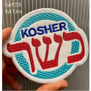 Puede incluir: Parche blanco bordado con un borde azul y turquesa. El parche presenta una menorá roja y la palabra "KOSHER" en letras azules.