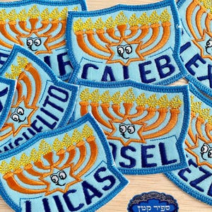 Puede incluir: Un conjunto de parches bordados azules y naranjas que representan una menorá con una cara de dibujos animados y un nombre debajo. Los nombres en los parches son: 'Caleb', 'Lucas', 'Eli', 'Josel' y 'Izzy'.