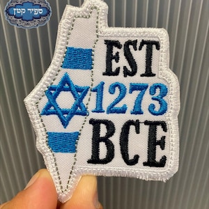 May include: A white embroidered patch shaped like the map of Israel, featuring a blue Star of David, blue horizontal stripes, and the black embroidered text EST 1273 BCE. The patch has a white stitched border and a white fabric base.