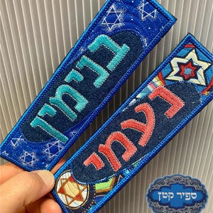 May include: Two embroidered blue fabric bookmarks with Hebrew text in teal and red. The bookmarks feature Star of David and other decorative patterns. One is held by a hand. A blue and silver emblem is in the lower right corner.