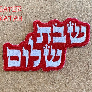 Puede incluir: Parche rojo bordado con letras hebreas blancas que se leen "Sapir Katan" en inglés. El parche es un símbolo judío tradicional.