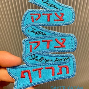 Puede incluir: Un parche bordado azul con letras hebreas rojas y las palabras en inglés "Justice" y "Shall you pursue". El parche tiene un diseño de cinta. El nombre "SAPIR KATAN" está en la parte inferior.