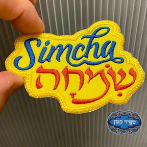 Könnte beinhalten: Gestickter gelber Aufnäher mit dem Wort "Simcha" in blauer Schrift über rotem hebräischen Text. Der Aufnäher hat einen dekorativen Rand und wird von einer Hand gehalten. Ein kleiner blau-weißer Aufkleber befindet sich in der unteren rechten Ecke.