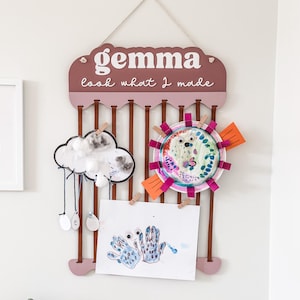 May include: A wooden display board with the name "gemma" and the phrase "look what I made". It features brown ribbons and wooden clips holding children's artwork, including a cloud, a sun, and handprint art.