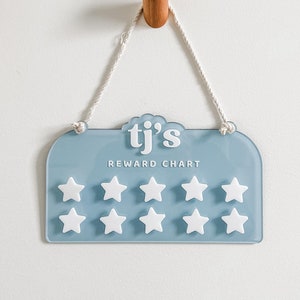 May include: A light blue reward chart with white star-shaped reward markers. The chart is labeled "tj's REWARD CHART" and hangs from a wooden peg with a white rope. The background is a plain white wall.