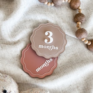 May include: Two scalloped-edge milestone markers in shades of brown and rose. The top marker reads "3 months" in white script. A wooden bead garland and a stuffed animal are also visible.