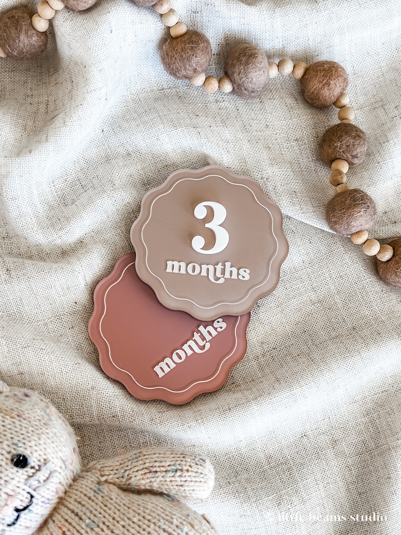 May include: Two scalloped-edge milestone markers in shades of brown and rose. The top marker reads "3 months" in white script. A wooden bead garland and a stuffed animal are also visible.