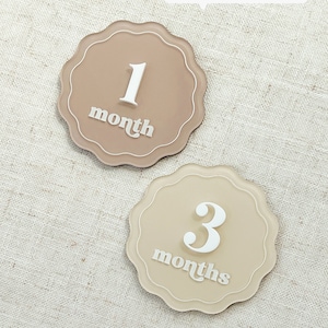 May include: Two beige, scalloped-edge milestone markers. One marker reads "1 month" in white script, the other reads "3 months". The text "DOUBLE SIDED ENGRAVED WITH 'MONTH' & 'MONTHS'" is above the markers.