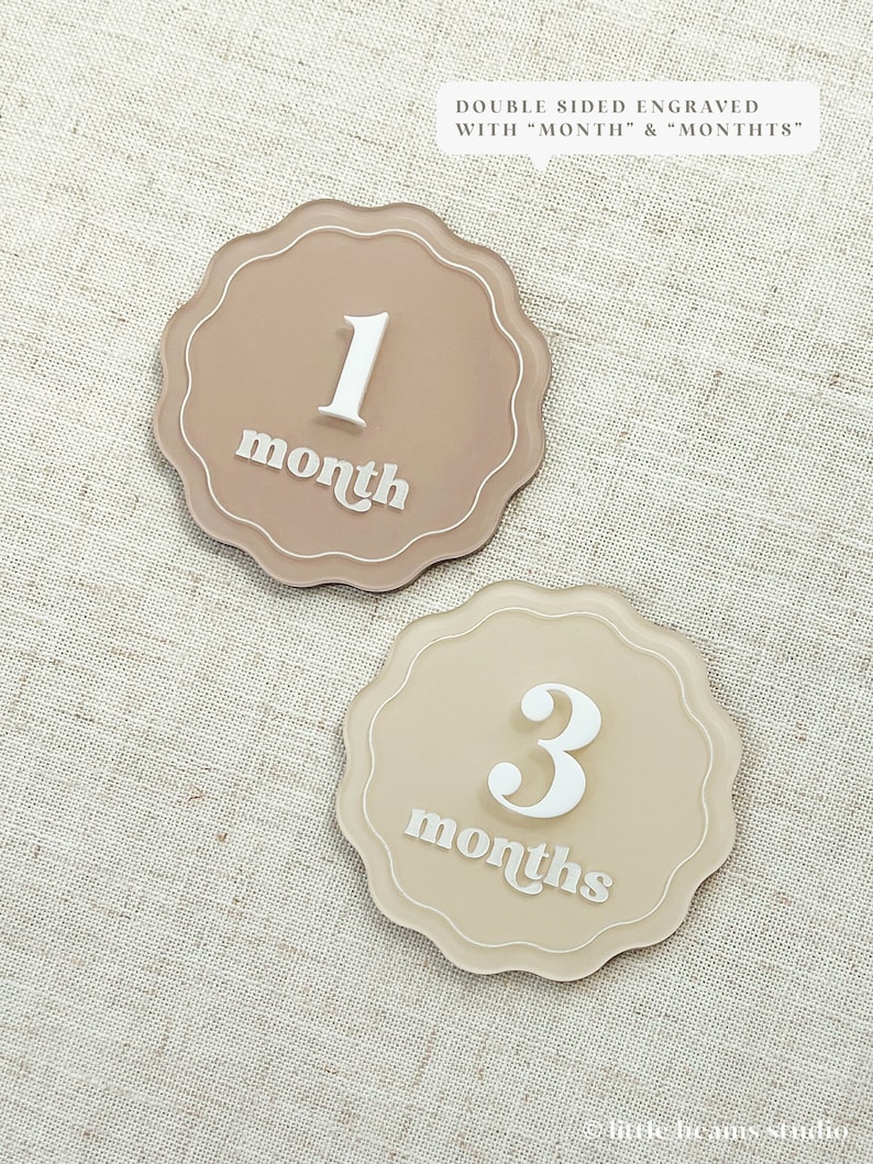 May include: Two beige, scalloped-edge milestone markers. One marker reads "1 month" in white script, the other reads "3 months". The text "DOUBLE SIDED ENGRAVED WITH 'MONTH' & 'MONTHS'" is above the markers.