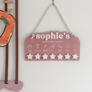 May include: A pink bedtime chart, labelled "Sophie's", with icons for bedtime routines. Includes spaces for stars as rewards. The chart is suspended by a rope and is designed for a child's bedroom.