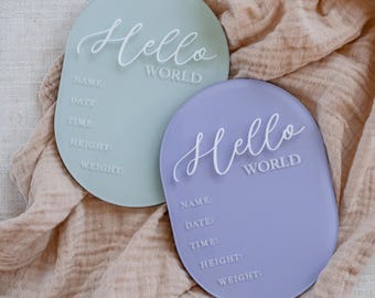 May include: Two oval-shaped baby announcement signs. One is light green, the other is lavender. Both have the words "Hello World" in cursive and spaces for name, date, time, height, and weight.