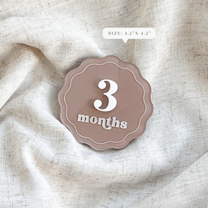 May include: A brown, scalloped-edge milestone marker with the number "3" and the word "months" in white. The marker is 4.5" x 4.5" and is set against a textured, neutral-toned fabric background. The marker is designed for baby photos.