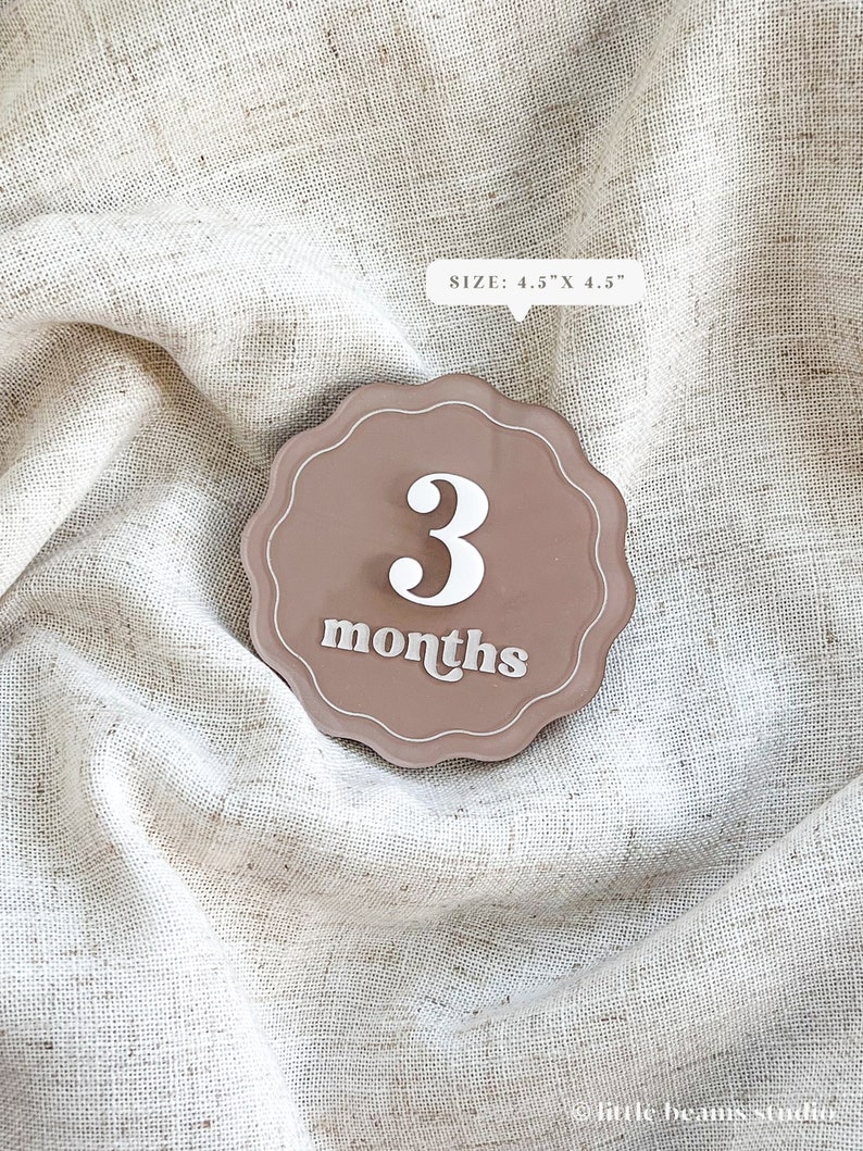 May include: A brown, scalloped-edge milestone marker with the number "3" and the word "months" in white. The marker is 4.5" x 4.5" and is set against a textured, neutral-toned fabric background. The marker is designed for baby photos.