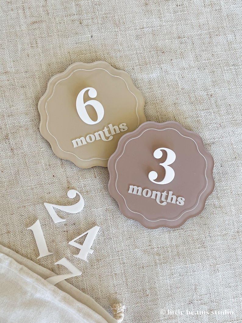 May include: Two scalloped-edge milestone markers, one tan and one brown, with the numbers 6 and 3, and the word "months" in white. Additional white numbers 1, 2, and 4 are also present. The items are on a textured beige surface.